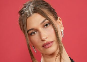 Hailey Bieber wears soft glam makeup and a slicked-back updo with face-framing pieces against a red backdrop