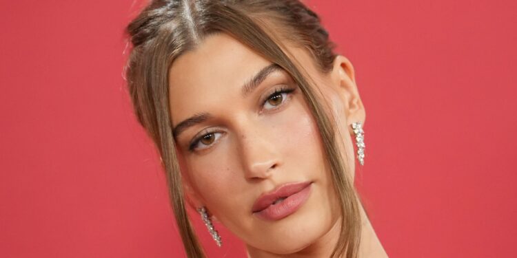 Hailey Bieber wears soft glam makeup and a slicked-back updo with face-framing pieces against a red backdrop