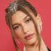 Hailey Bieber wears soft glam makeup and a slicked-back updo with face-framing pieces against a red backdrop