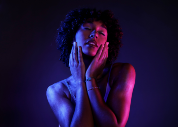 Close-up of a woman with curly hair, eyes closed, holding both hands to her cheeks under blue and pink lighting.