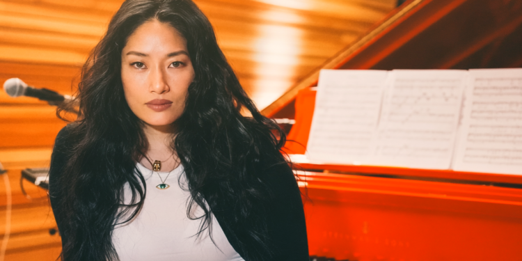 Chloe Flower seated in front of a red grand piano with sheet music, wearing a black jacket over a white top.