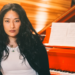 Chloe Flower seated in front of a red grand piano with sheet music, wearing a black jacket over a white top.